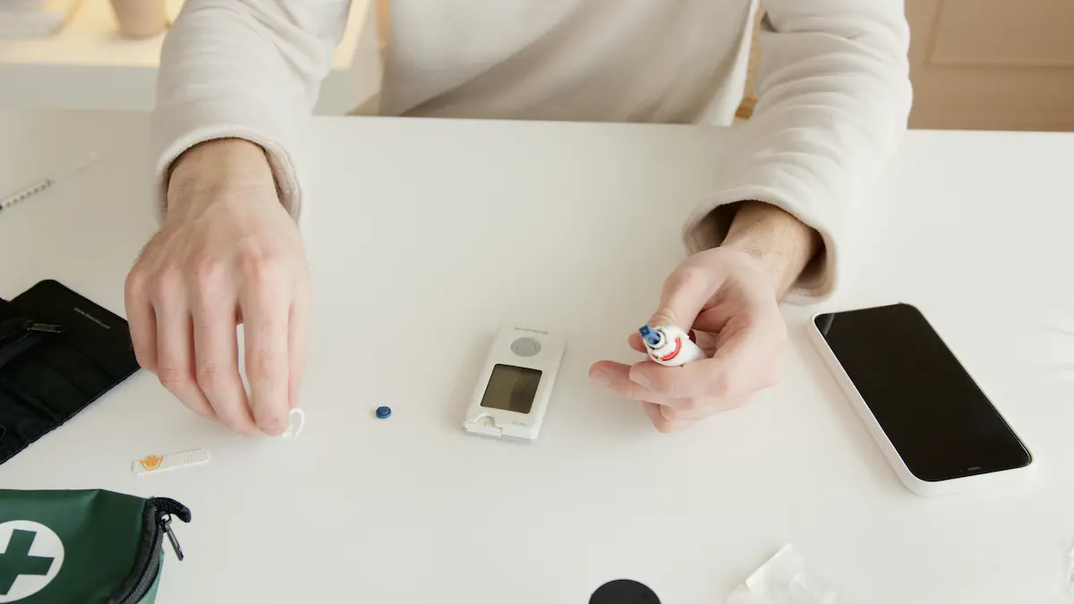 Person preparing a glucose test with a glucometer, lancet device, and medical kit on a white table